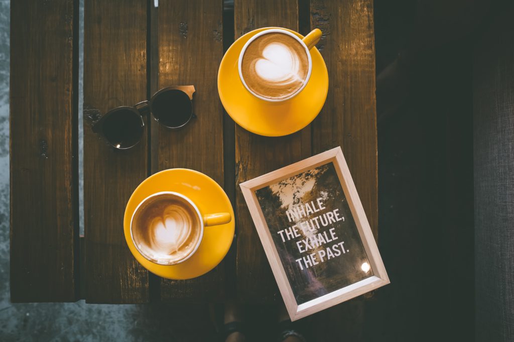 A wooden table with two cups of coffee served on small yellow plates, sunglasses, and an inspiring framed picture laying on the table.